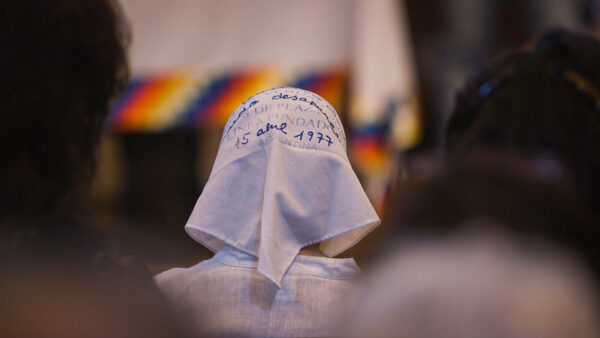 A person wearing a white headscarf with handwritten blue text sits among others in a solemn gathering.