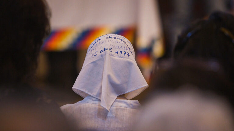 A person wearing a white headscarf with handwritten blue text sits among others in a solemn gathering.