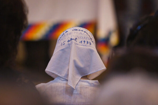 A person wearing a white headscarf with handwritten text, including the date "15 abril 1977," sits among a group of people; a colorful blur appears in the background.