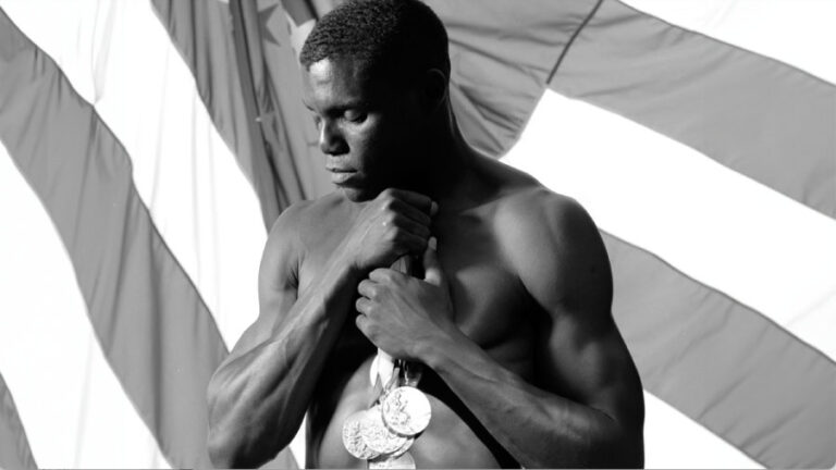 A shirtless athlete showcases a muscular physique, holding a collection of medals close to his chest as he stands in front of a large American flag. The artistic black-and-white composition, with strong light and shadow, emphasizes strength, achievement, and national pride, likely celebrating a record of Olympic or world-class triumphs.
