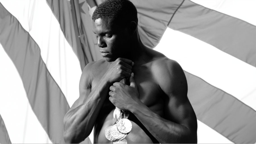 A shirtless athlete showcases a muscular physique, holding a collection of medals close to his chest as he stands in front of a large American flag. The artistic black-and-white composition, with strong light and shadow, emphasizes strength, achievement, and national pride, likely celebrating a record of Olympic or world-class triumphs.