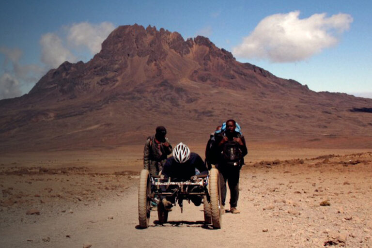 Three people, including one using a handcycle, travel on a dirt path toward a rocky mountain under a partly cloudy sky.