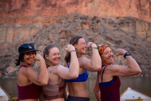 Four women stand side by side in a canyon, flexing their arms to show off their muscles. They wear athletic tops and sports watches or wristbands. The rocky canyon wall and river in the background suggest an outdoor adventure or rafting trip. The scene conveys camaraderie, strength, and empowerment among the group.