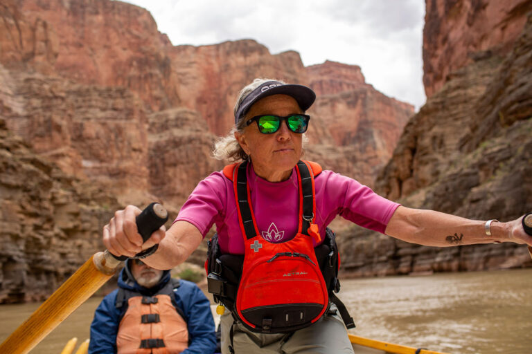A person in a bright pink shirt and red life jacket is rowing a boat through a dramatic canyon landscape. Towering reddish rock walls rise on either side of the muddy river, and another rower wearing a blue jacket and tan life vest sits behind. The scene conveys a sense of adventure and teamwork among the striking natural formations.