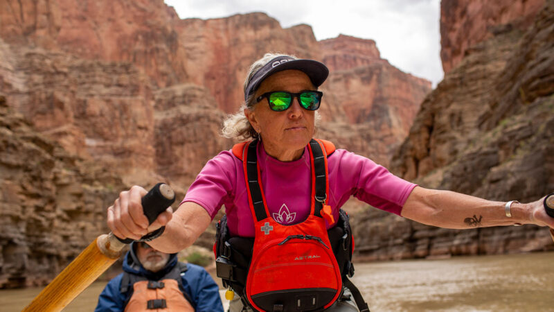 A person in a bright pink shirt and red life jacket is rowing a boat through a dramatic canyon landscape. Towering reddish rock walls rise on either side of the muddy river, and another rower wearing a blue jacket and tan life vest sits behind. The scene conveys a sense of adventure and teamwork among the striking natural formations.