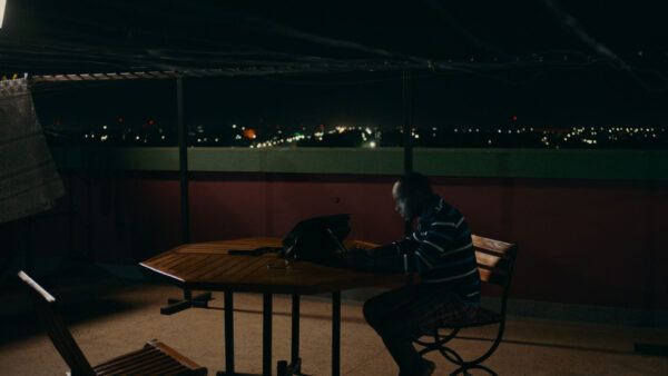 A person sits at a wooden table on a rooftop at night, working on a laptop. City lights twinkle in the distance, creating a serene, focused atmosphere.