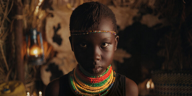 A person wearing colorful bead necklaces, sitting indoors with warm ambient lighting, looking at the camera intently.