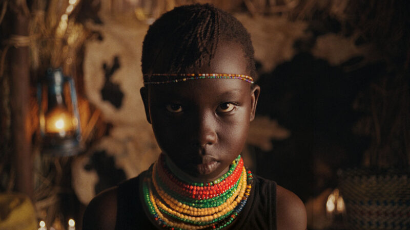 A person wearing colorful bead necklaces, sitting indoors with warm ambient lighting, looking at the camera intently.