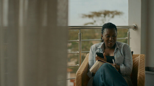 A woman sits on a balcony in a wicker chair, smiling at her phone with a notebook resting on her lap.