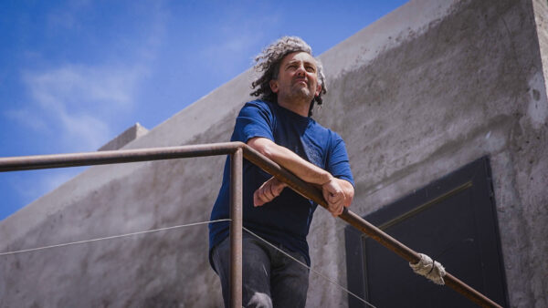 A man with curly hair leans on a railing, gazing thoughtfully into the distance against a clear blue sky and concrete building backdrop.