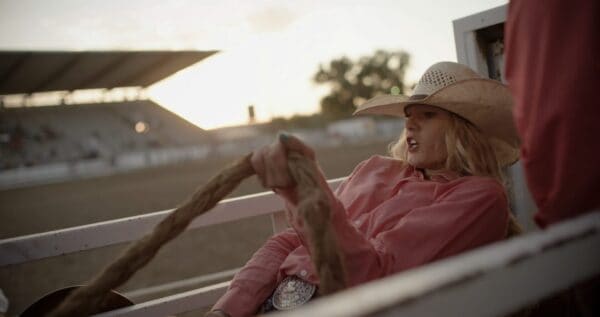 A woman wearing a cowboy hat and pink shirt in a horse racing track during sunset.