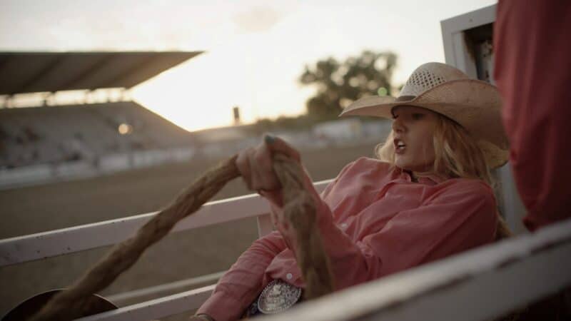 A woman wearing a cowboy hat and pink shirt in a horse racing track during sunset.