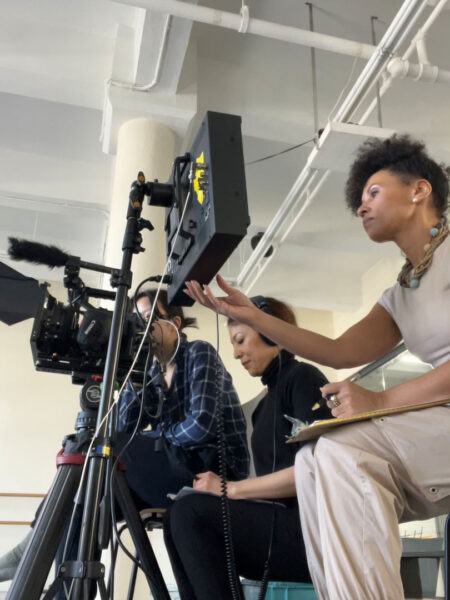 A small film crew is at work indoors, with a camera on a tripod capturing the scene and several people monitoring a large screen attached to the rig. One person sits nearby holding a notepad and gesturing toward the monitor, while another operates equipment and another wears headphones, reflecting a collaborative, behind-the-scenes atmosphere on a film or documentary set.