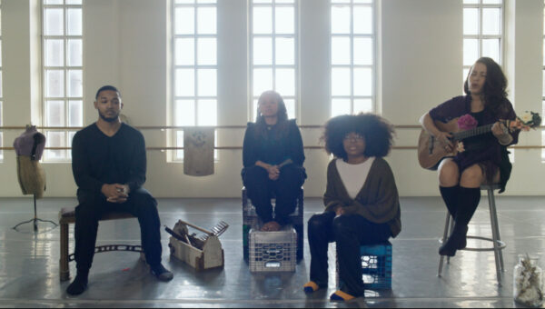 In a sunlit studio with tall windows, four people sit in a row—two on crates, one on a stool playing a guitar decorated with flowers, and one at the left on a standard chair. The group is surrounded by objects including fabric, crates, and a dress form, creating a creative and intimate setting that suggests rehearsal, performance, or a collaborative workshop.