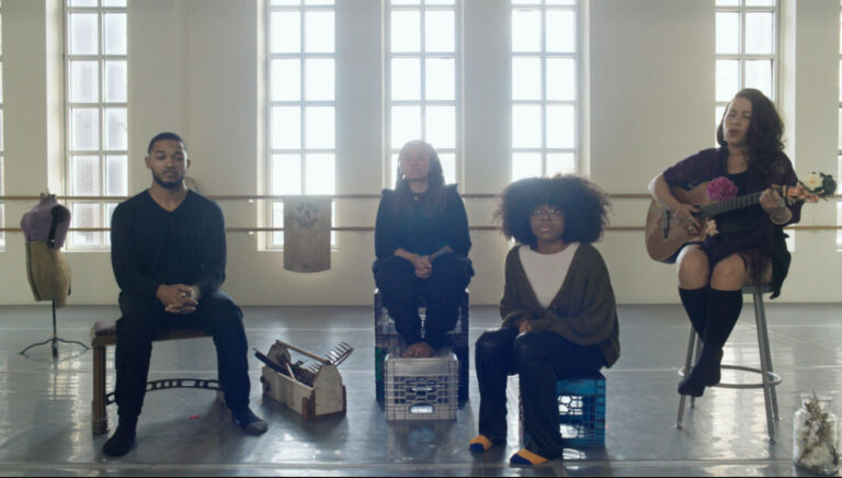 In a sunlit studio with tall windows, four people sit in a row—two on crates, one on a stool playing a guitar decorated with flowers, and one at the left on a standard chair. The group is surrounded by objects including fabric, crates, and a dress form, creating a creative and intimate setting that suggests rehearsal, performance, or a collaborative workshop.