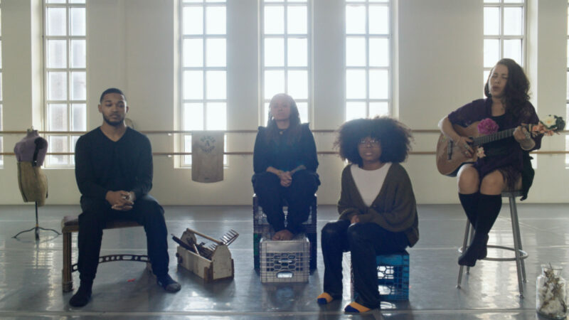 In a sunlit studio with tall windows, four people sit in a row—two on crates, one on a stool playing a guitar decorated with flowers, and one at the left on a standard chair. The group is surrounded by objects including fabric, crates, and a dress form, creating a creative and intimate setting that suggests rehearsal, performance, or a collaborative workshop.