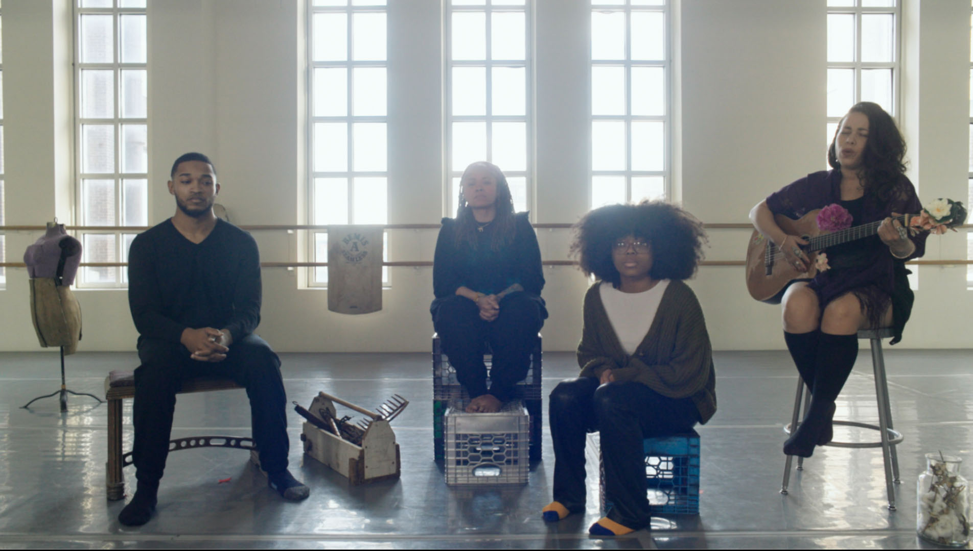 In a sunlit studio with tall windows, four people sit in a row—two on crates, one on a stool playing a guitar decorated with flowers, and one at the left on a standard chair. The group is surrounded by objects including fabric, crates, and a dress form, creating a creative and intimate setting that suggests rehearsal, performance, or a collaborative workshop.