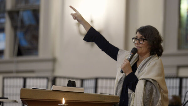 A person stands at a podium inside a well-lit, spacious room filled with soft light. They hold a microphone and gesture energetically with one hand raised, addressing an audience. An open book sits on the podium next to a lit candle, and the scene conveys a sense of ceremony, inspiration, or communal gathering.