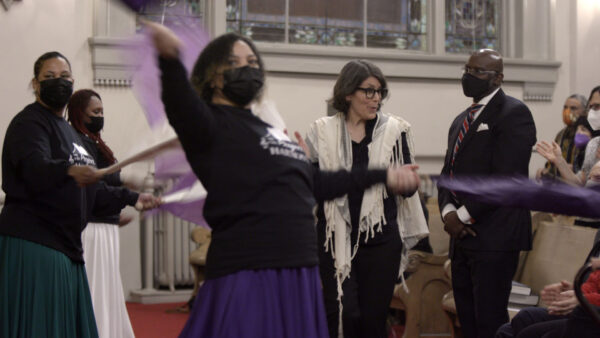 People participate in an expressive and energetic ceremony inside a church, surrounded by stained glass windows and pews. Some individuals wave purple and white fabric streamers, creating a sense of movement. Others, including a woman wearing a fringed shawl, stand attentively or watch from the congregation, contributing to a vibrant, communal atmosphere.