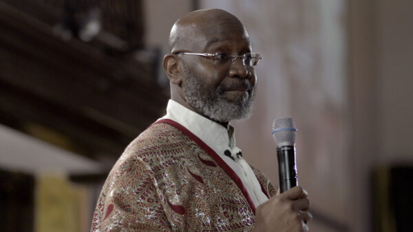 A person holds a microphone while speaking in a public setting, wearing a white shirt with an ornate, patterned shawl or jacket. The background is softly lit and out of focus, suggesting an indoor gathering such as a ceremony, service, or presentation.