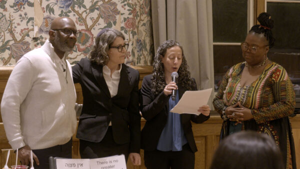Four people stand together indoors in front of floral wallpaper and curtains, dressed in formal or semi-formal attire. One person reads from a sheet while holding a microphone, two others listen closely, and the fourth stands with hands clasped. The group appears to be participating in a ceremony, speech, or presentation, creating an atmosphere of collaboration and respect.