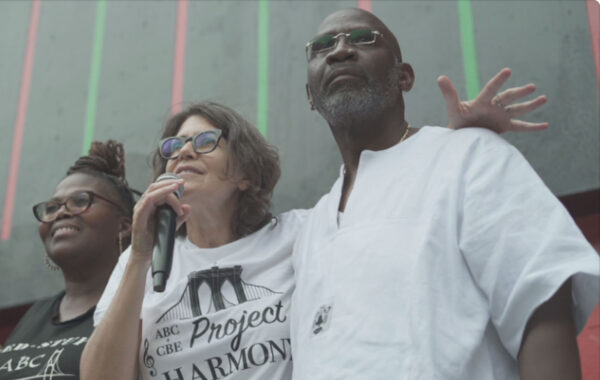 Three people stand together in front of a gray wall with vertical neon stripes, embracing with arms around each other. One person in the center holds a microphone and wears a white t-shirt with a "Project Harmony" logo featuring a stylized Brooklyn Bridge. The atmosphere is warm and communal, suggesting a moment of celebration or unity at a public event.