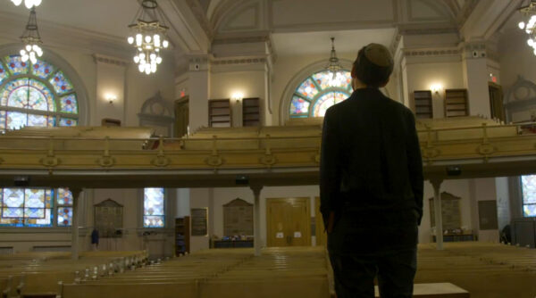 A person stands inside a spacious synagogue, looking up at the balcony seating with two large stained glass rose windows in the background. The interior features high ceilings, ornate light fixtures, wooden pews, and soft cream walls, creating a peaceful atmosphere filled with natural and artificial light.