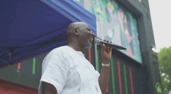 A person in a white shirt stands under a blue canopy, holding a microphone. Behind them is a brightly colored mural with abstract shapes and figures, while vertical neon stripes accent the wall. The outdoor event setting and confident posture suggest public speaking or performance.