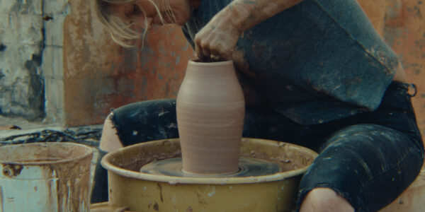 A person sits at a pottery wheel, shaping a tall clay vessel with both hands. Their clothes and arms are covered in clay, and containers filled with tools and materials surround them. The rustic, textured wall in the background adds to the artistic atmosphere of the workspace.