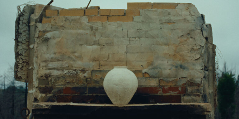 A single, large clay pot sits on a wooden shelf in front of a weathered, partially crumbled brick and plaster wall. The outdoor scene feels rustic and quiet, with muted colors and rough textures highlighting the handmade pottery against the aged structure.