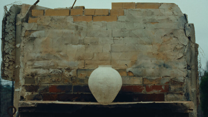 A single, large clay pot sits on a wooden shelf in front of a weathered, partially crumbled brick and plaster wall. The outdoor scene feels rustic and quiet, with muted colors and rough textures highlighting the handmade pottery against the aged structure.