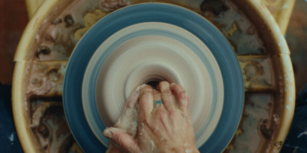 An overhead view of a pottery wheel spinning as hands shape wet clay into a vessel. The wheel and surrounding area are splattered with clay, capturing the dynamic motion and tactile process of ceramics making.