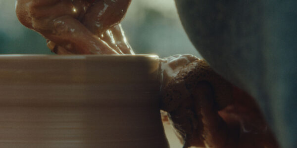 A close-up of hands working with wet clay on a pottery wheel, gently shaping the rim of a vessel. The person's fingers are covered in clay and they use a natural sponge, capturing the artistry and tactile details of ceramic creation. Warm light highlights the textures of the clay and hands.