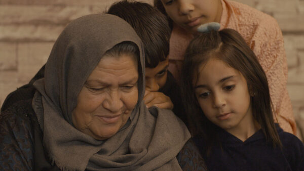 Three children and an adult sit closely together, displaying a sense of warmth and intimacy. The adult wears a gray headscarf and dark patterned clothing, while the children gather near, partially leaning on one another. Their expressions and posture suggest comfort and family affection, set against a softly lit brick-patterned background.