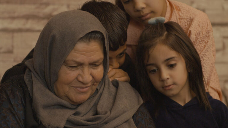 Three children and an adult sit closely together, displaying a sense of warmth and intimacy. The adult wears a gray headscarf and dark patterned clothing, while the children gather near, partially leaning on one another. Their expressions and posture suggest comfort and family affection, set against a softly lit brick-patterned background.