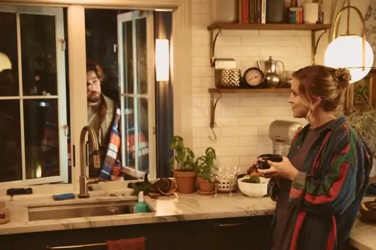 Woman in colorful striped robe smiling at man outside kitchen window during evening, warm indoor lighting, cozy home setting with plants and kitchenware.