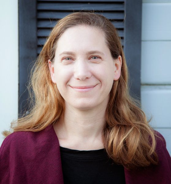 A woman with long light brown hair is standing outside in front of a white wall with a dark shutter, wearing a maroon sweater over a black top, and smiling at the camera.