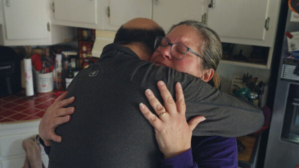 A person embraces another individual tightly in a cozy kitchen. The hand of one person, featuring a gold ring, is gently resting on the other's back. The surrounding area includes kitchen cabinets, red tiled counters, and various utensils, creating a warm, domestic atmosphere.