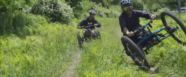 Two mountain bikers riding downhill on a grassy trail surrounded by green trees and bushes, enjoying outdoor adventure and nature.