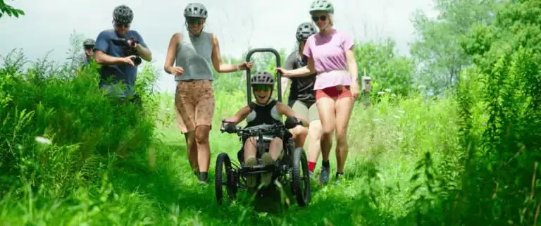Group of young people riding bikes and walking on a lush green trail in a park during daytime, enjoying outdoor activity and nature.