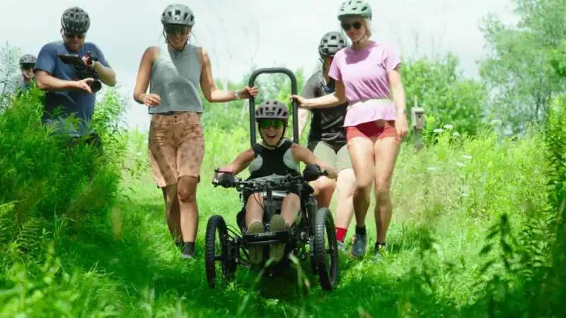 Group of young people riding bikes and walking on a lush green trail in a park during daytime, enjoying outdoor activity and nature.