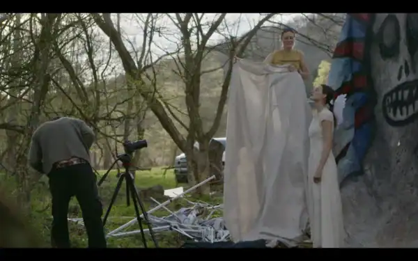 Filmmakers capture a scene with two women in costume, one holding a fabric backdrop, outdoors among trees, with a camera on a tripod and crew members setting up.