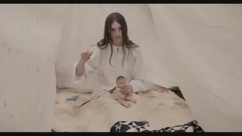 A woman with long dark hair sitting on a bed holding a baby, surrounded by white fabric drapes, in a minimalist setting for a film or art project.