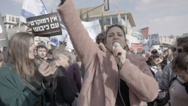 A crowd gathers outdoors for a protest or demonstration, with people holding signs in Hebrew and waving both Israeli and Palestinian flags. The central figure in a light pink coat raises a fist and speaks passionately into a microphone, while others clap and display supportive gestures. Buildings and a clear sky fill the background, creating an atmosphere of collective action and activism in a public space.