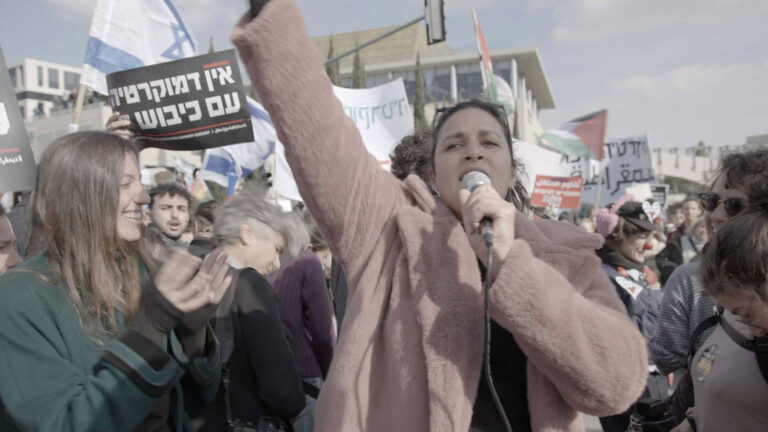 A crowd gathers outdoors for a protest or demonstration, with people holding signs in Hebrew and waving both Israeli and Palestinian flags. The central figure in a light pink coat raises a fist and speaks passionately into a microphone, while others clap and display supportive gestures. Buildings and a clear sky fill the background, creating an atmosphere of collective action and activism in a public space.