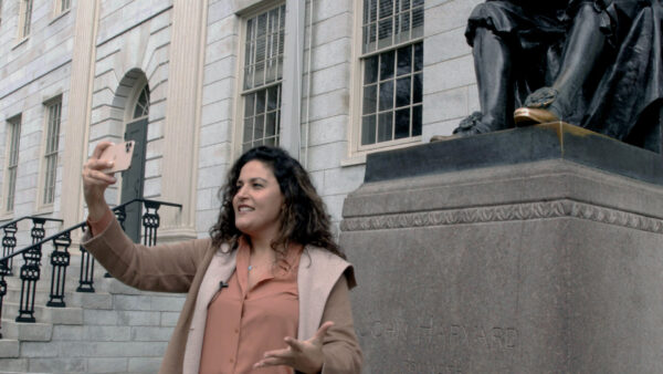 A person stands outside a historic stone building, holding up a smartphone to take a selfie in front of a large bronze statue seated on a stone base. The engraved text on the base reads "John Harvard," identifying the famous statue located at Harvard University. The individual wears a light brown coat over a salmon shirt, capturing a moment of their visit on a campus known for its iconic architecture and academic reputation.