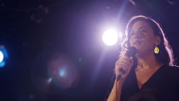 A person stands on stage holding a microphone, illuminated by striking stage lighting that creates dramatic lens flares in the image. The individual wears a dark V-neck outfit, a delicate chain necklace, and bold gold earrings shaped with script-like designs. The setting conveys the atmosphere of a live performance with a focus on powerful expression and presence.