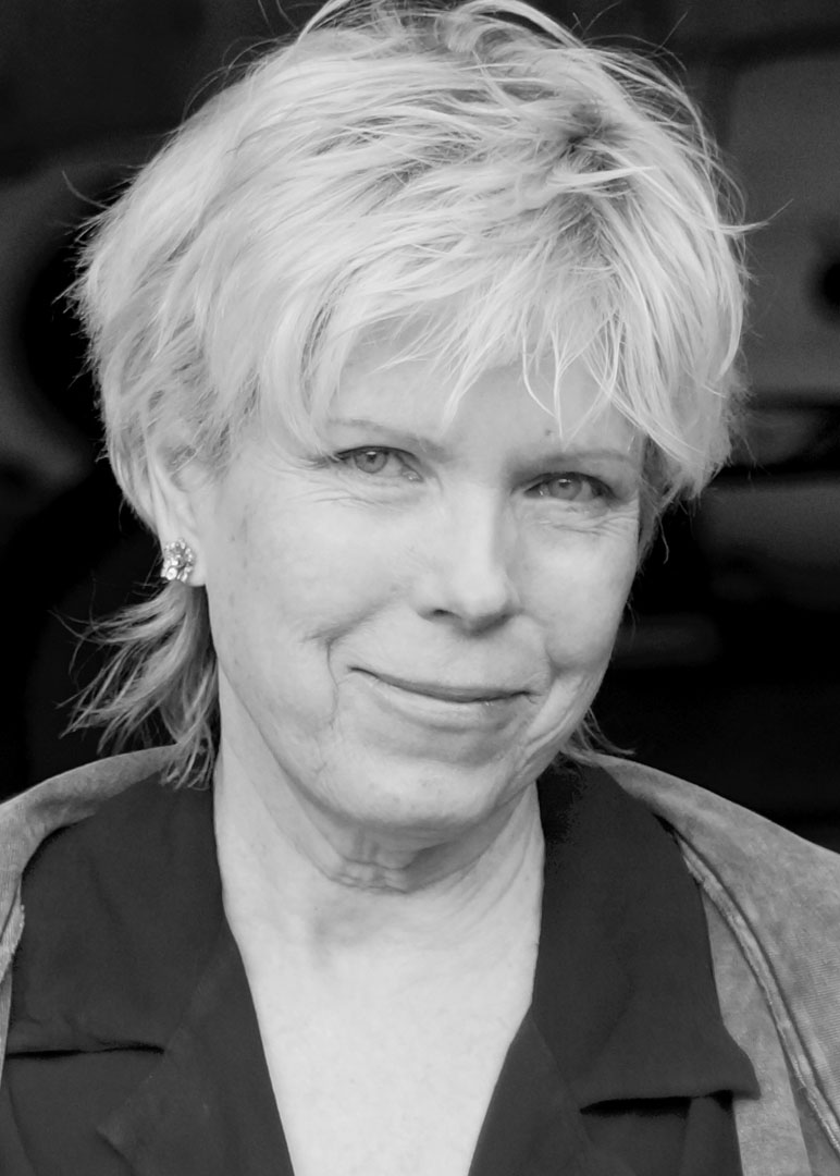Black and white headshot of a woman with light-colored, short, tousled hair, small earrings, and a soft smile, wearing a dark collared shirt and light jacket.