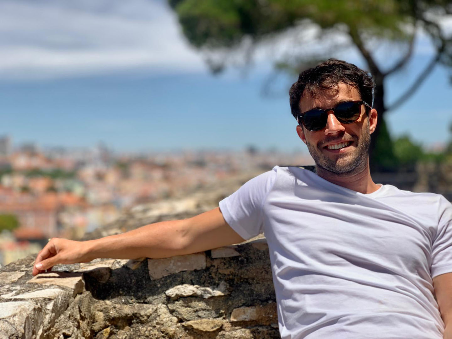 A man relaxing outdoors on a sunny day, leaning back against a stone wall. He is wearing a white t-shirt and sunglasses, smiling warmly at the camera. The background is brightly blurred but suggests a scenic overlook or cityscape with a clear sky and greenery, creating a casual and cheerful atmosphere.
