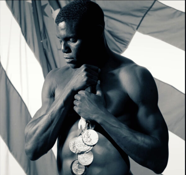 A shirtless athlete showcases a muscular physique, holding a collection of medals close to his chest as he stands in front of a large American flag. The artistic black-and-white composition, with strong light and shadow, emphasizes strength, achievement, and national pride, likely celebrating a record of Olympic or world-class triumphs.